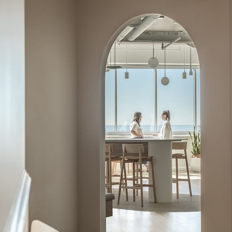Two women in white attire engage in conversation at a modern, minimalist wooden table by a large window overlooking the ocean