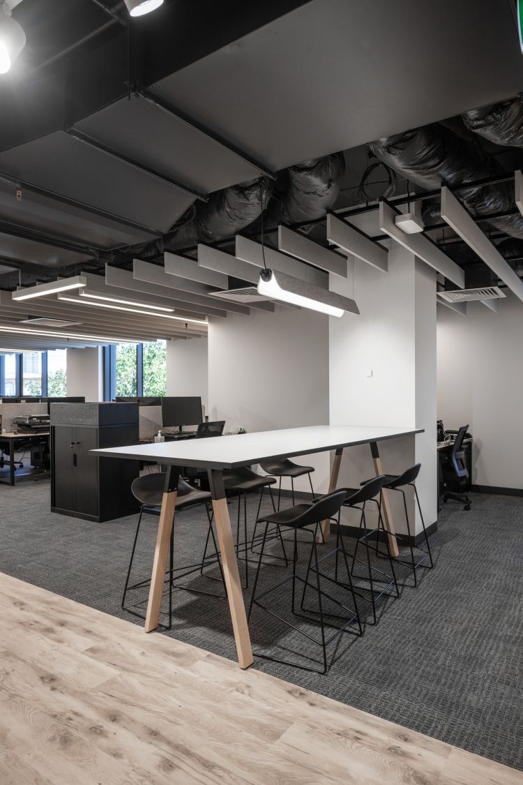  A modern office space with sleek design elements, featuring tiered overhead lighting, organized workstations with black chairs, and a carpeted floor transitioning to wooden flooring.