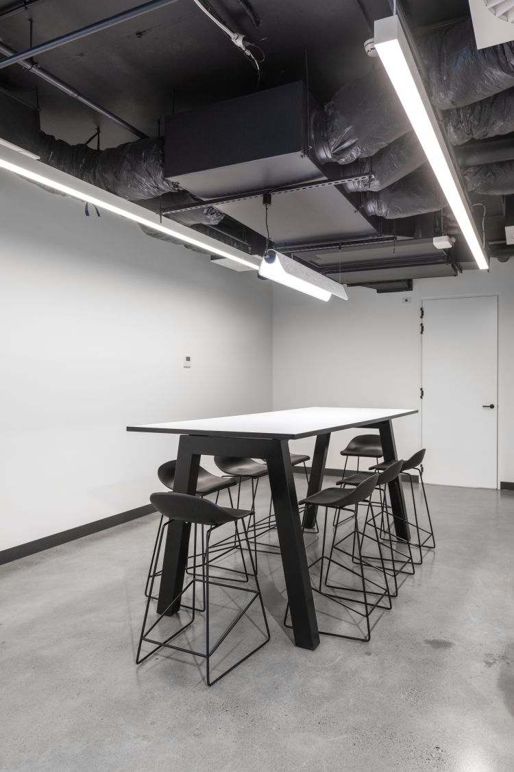 A minimalist office space with a sleek white table paired with black high stools, set against a polished concrete floor. 