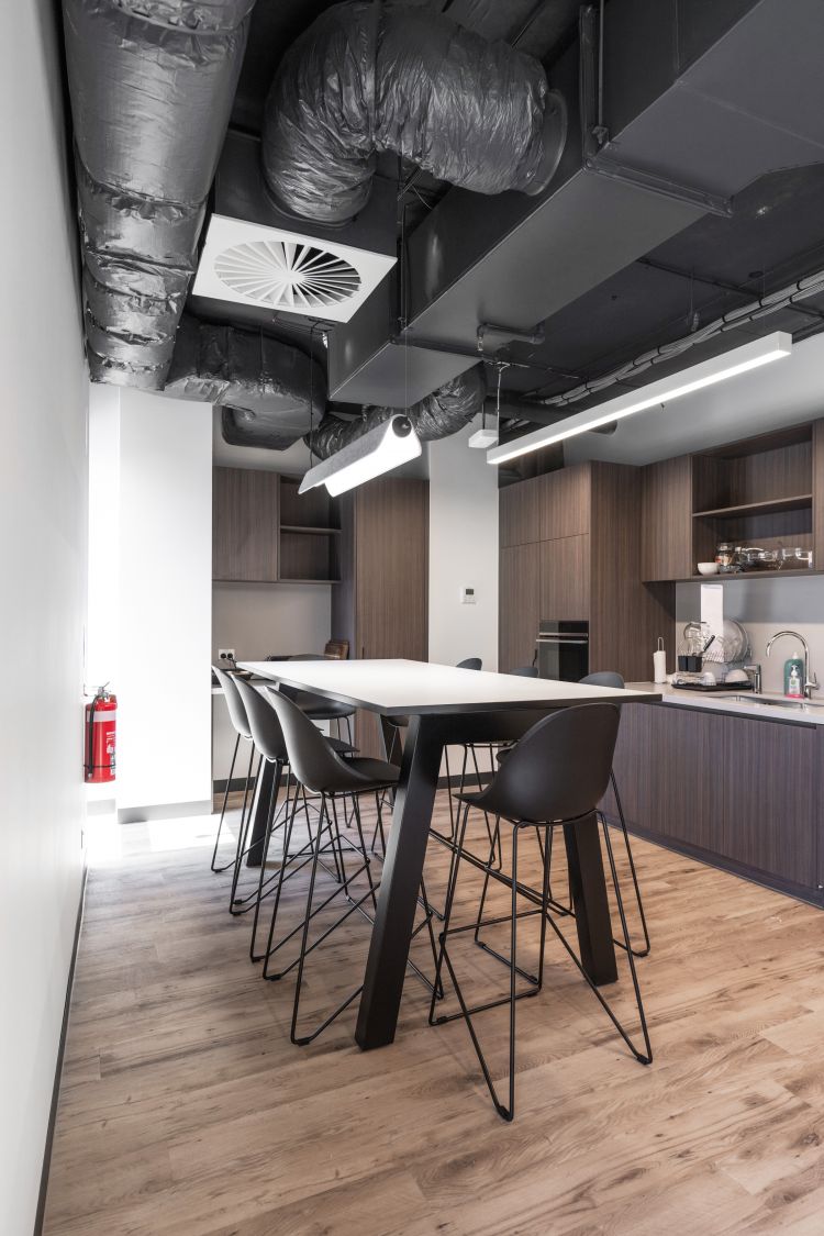 An office kitchen space with a table surrounded by black high stools, providing a casual dining or meeting area.