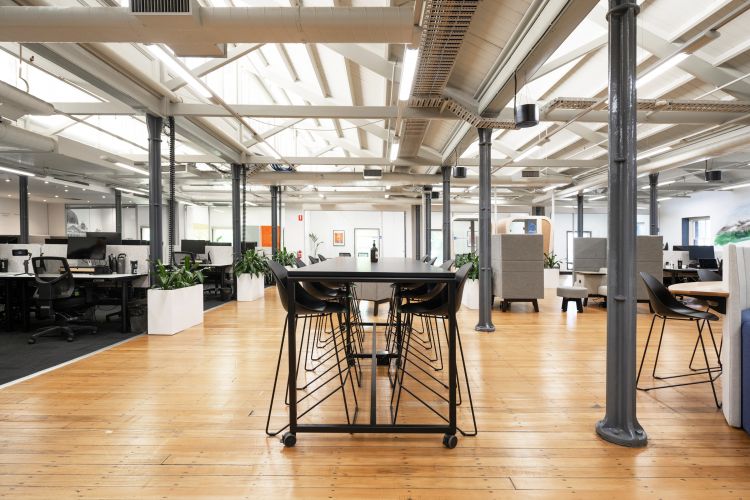 A spacious modern office with an open floor plan, featuring wooden flooring, black desks with computer setups, and exposed ceiling beams.