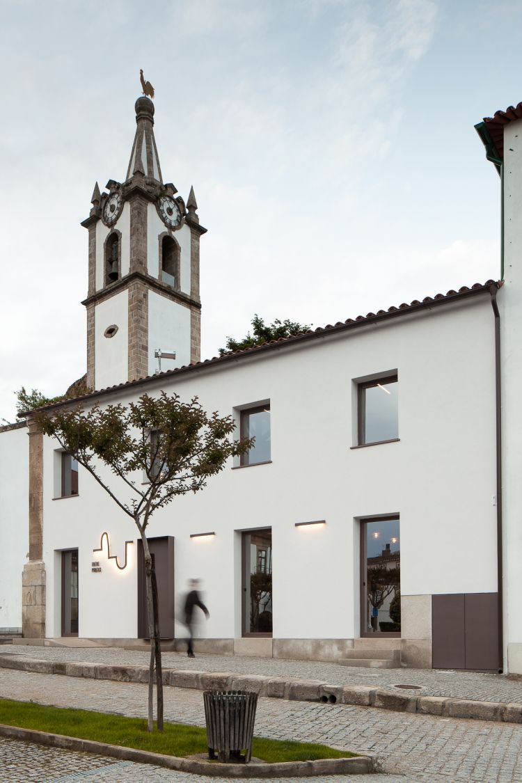 Historic stone bell tower with a clock and weather vane, adjacent to a modern white building with squared windows.