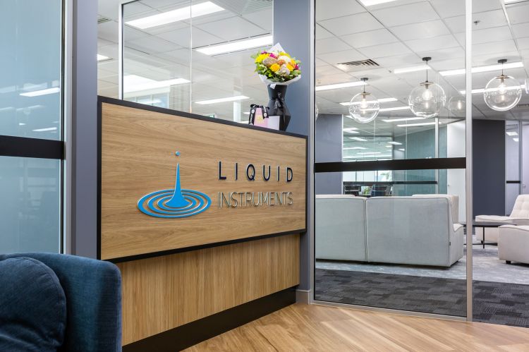 Modern office interior featuring a wooden reception desk with the "Liquid Instruments" logo and emblem illuminated in blue.
