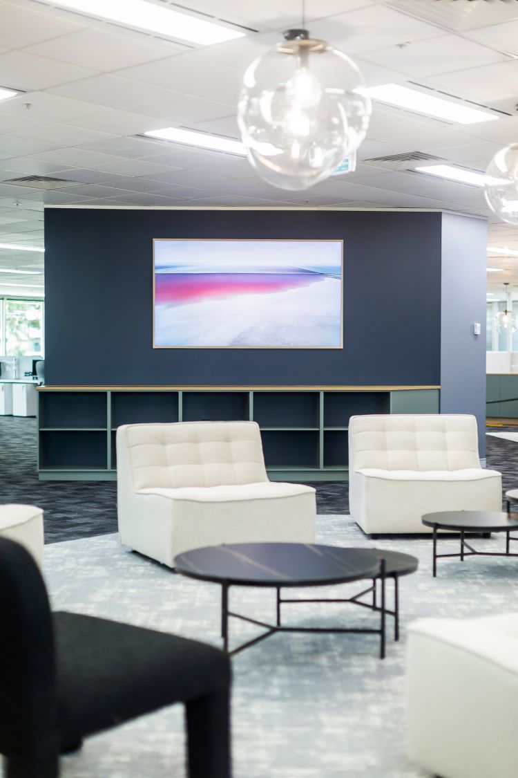 Modern office lounge area with a striking blue accent wall showcasing a large framed landscape artwork depicting serene shades of blue, pink, and white.