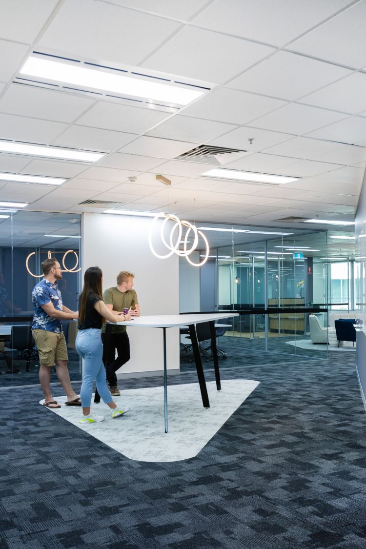 Modern office interior with a group of three individuals engaged in a casual conversation around a white standing table.