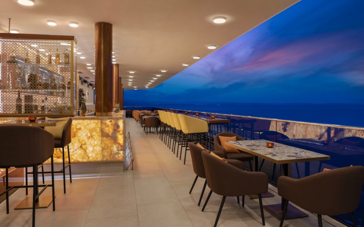 Elegant lounge area with a backlit bar, decorative wine display, and a row of golden-hued chairs facing large windows showcasing a serene twilight seascape.