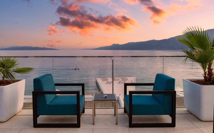 Tranquil balcony setting with turquoise armchairs and a small table, overlooking an infinity pool that blends into a serene seascape beneath a fiery sunset sky with mountains in the distance. 