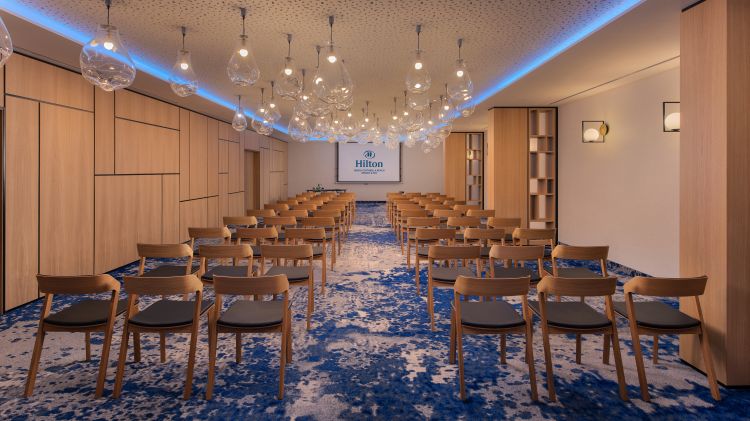 Modern conference room in a Hilton hotel featuring rows of wooden chairs with gray cushions, intricate blue carpeting, and unique glass pendant lights.