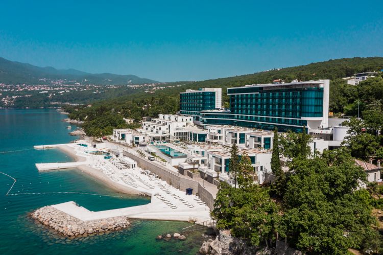 Aerial view of a coastal luxury resort with modern white buildings and a turquoise glass-facade hotel.