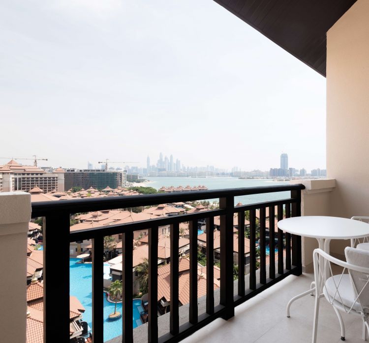 Spacious balcony view overlooking a resort with brown-tiled rooftops, a blue swimming pool, and palm trees.