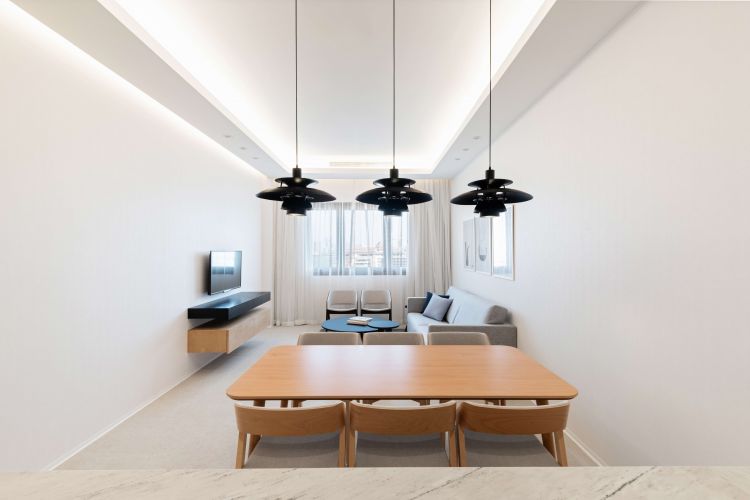 Contemporary living space with a prominent linear lighting feature on the ceiling, three hanging black pendant lights, a wooden dining table accompanied by matching chairs,