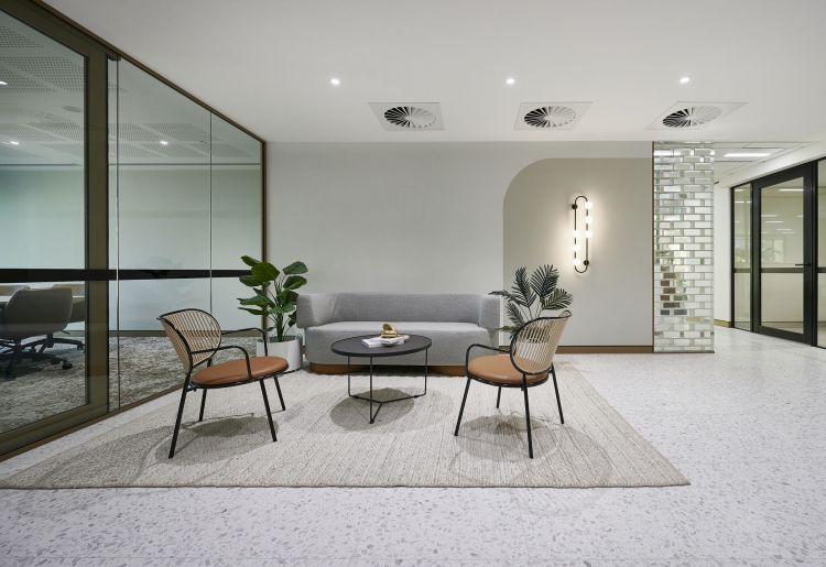 A modern office lounge area featuring a light gray sofa, two stylish chairs with wooden accents, a round coffee table