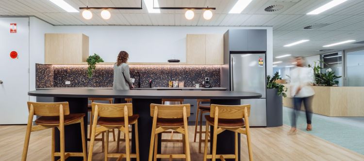 Contemporary office kitchen area surrounded by wooden stools, pendant lights, and plants.