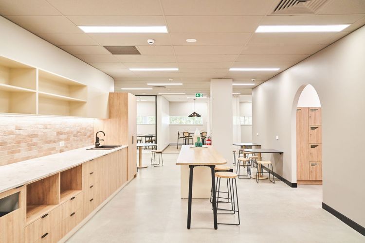 Modern office kitchen area with wooden countertops, shelves, and furniture, adjacent to a spacious dining space with tables, chairs, and an arched wooden door