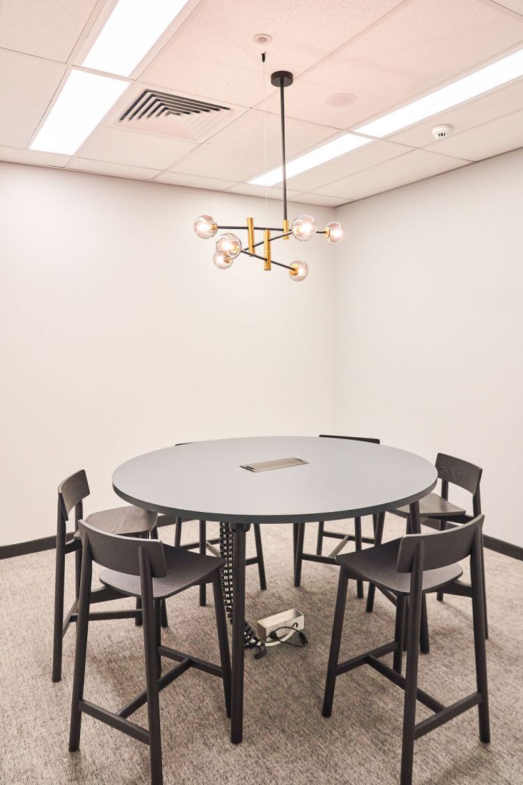 Minimalist conference room with a gray round table, dark chairs, and an overhead gold-toned chandelier with spherical bulbs.