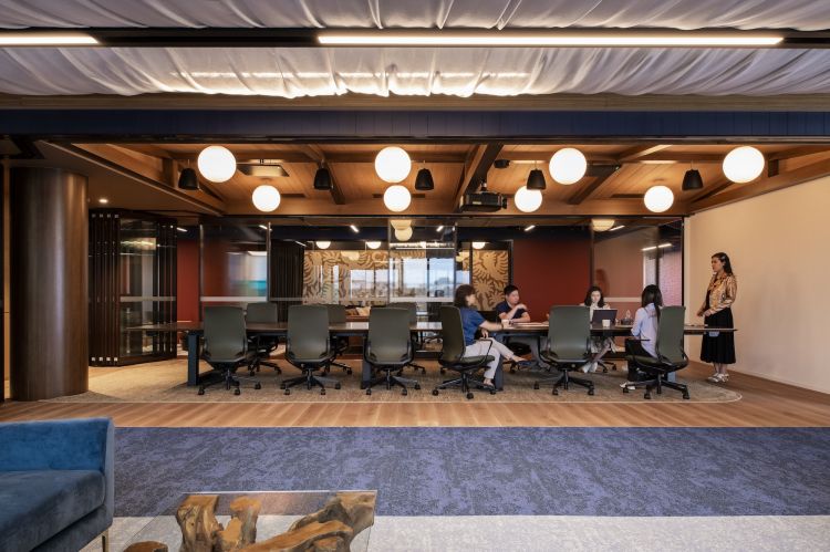 An office interior with a contemporary design, featuring a spacious meeting area where people are gathered around a long table with green chairs.