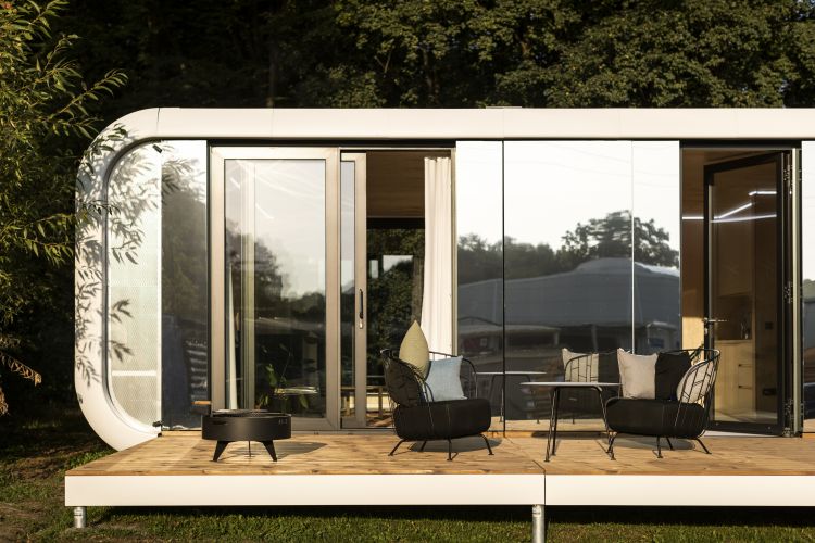 An exterior view of a modern modular home with curved corners, expansive glass windows, and a sliding door that opens onto a wooden deck with a fire pit and two metal frame chairs with cushions