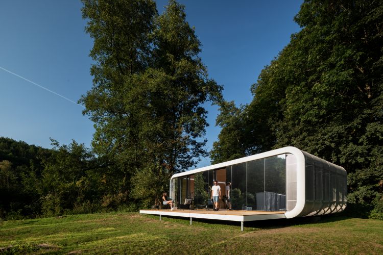 A modern, curved modular home with floor-to-ceiling windows, situated in a lush green clearing surrounded by tall trees