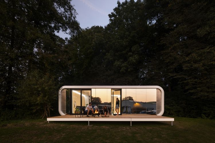 An evening scene of a modern, streamlined modular home with curved edges and full-length windows, warmly lit from within
