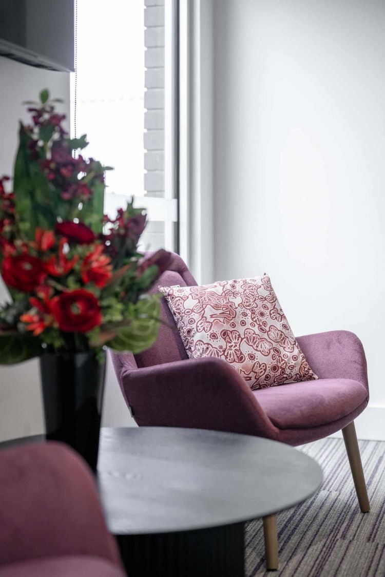 A contemporary purple armchair with a white and red patterned cushion, positioned next to the Mimi table with a vase of red flowers, against the backdrop of a bright window and white wall