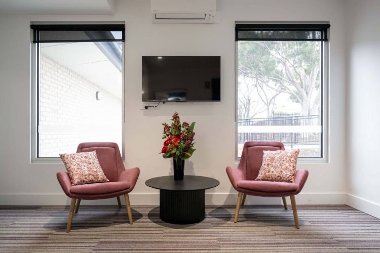 Two matching purple armchairs with patterned cushions on either side of a round black table with a vase of red flowers, set in a room with large windows