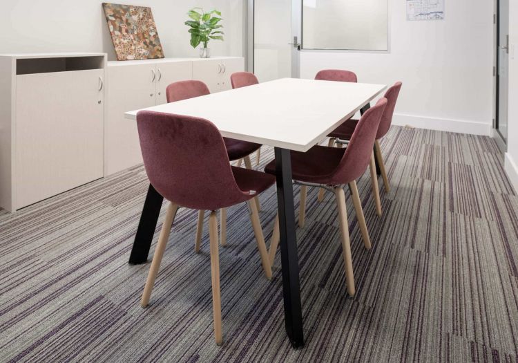 A modern meeting room featuring a white rectangular table with black legs, surrounded by four purple chairs with wooden legs
