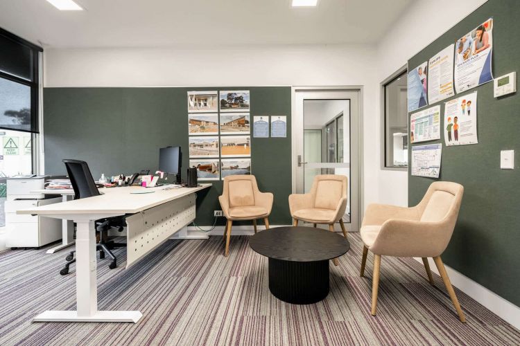 An office space with a large desk and computer, flanked by two beige armchairs and the Mimi coffee table. The room features a green accent wall with various posters and a glass door on the right.