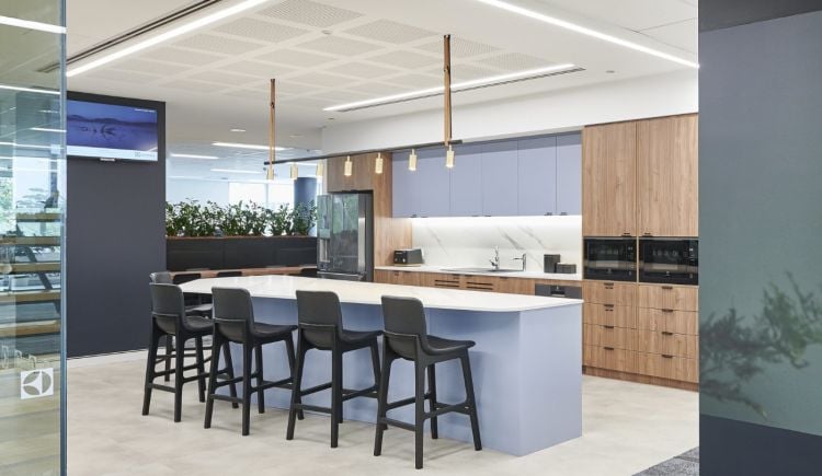 A modern office kitchenette with a central island and bar stools, wooden cabinetry, built-in appliances, and pendant lights, adjacent to a meeting area with a large table and chairs.