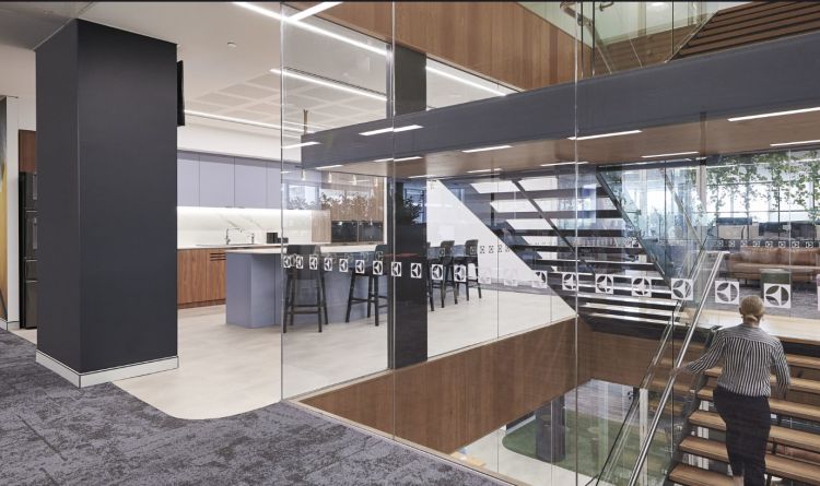 An office interior viewed through a glass partition, featuring a sleek kitchen area with bar stools and wooden accents, a staircase with glass railings leading to another level