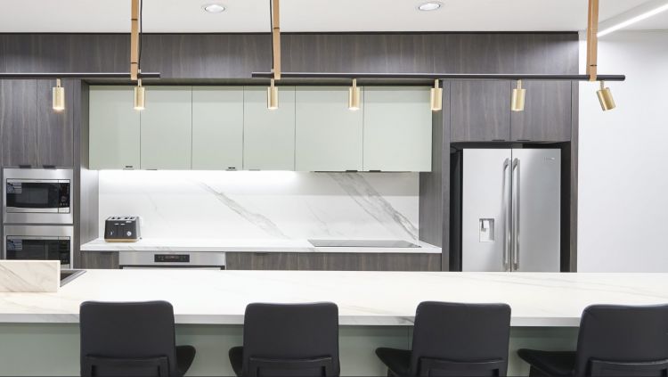 A sleek modern office kitchen with dark cabinetry, marble countertops, gold pendant lighting, and high-end appliances, including a stainless steel refrigerator and built-in ovens.