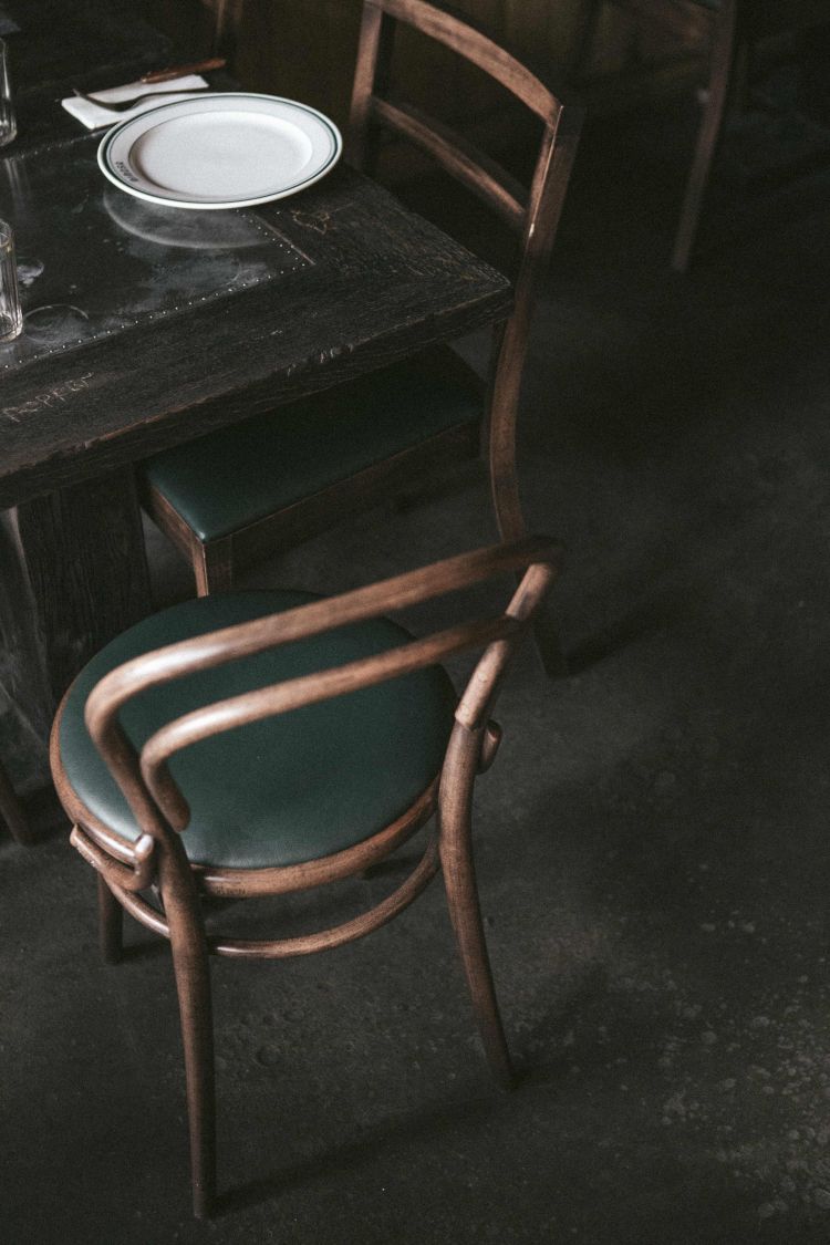 An old-fashioned wooden table with a set white plate, accompanied by a matching wooden chair with a green cushion, evoking a rustic and cozy dining atmosphere.