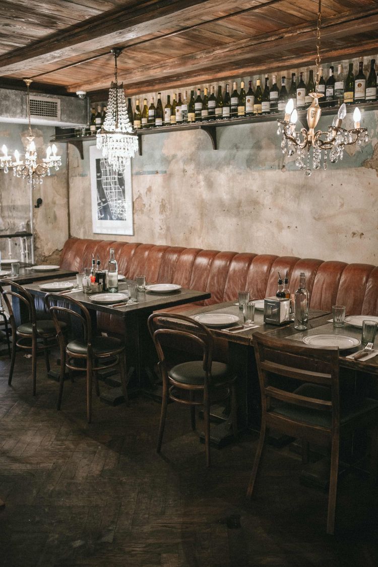 A rustic restaurant interior with a long leather bench, wooden tables and chairs, and crystal chandeliers. Wine bottles line a shelf above, contributing to the vintage atmosphere.