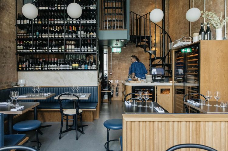 A cozy restaurant interior with a wine shelf, a spiral staircase, a view of the kitchen counter, and neatly arranged dining tables with blue banquettes.