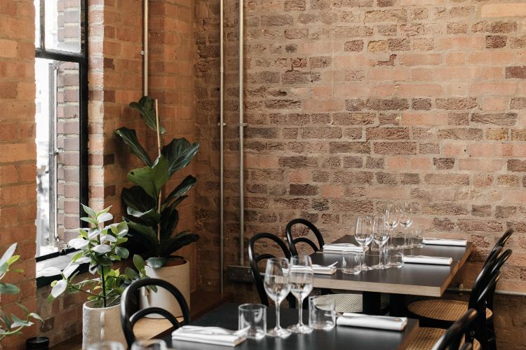 A rustic dining corner with exposed brick walls, potted plants, and tables set with wine glasses, adjacent to a window letting in natural light.
