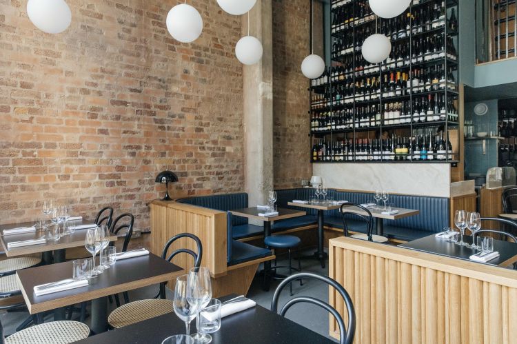Interior of a modern restaurant with exposed brick walls, spherical pendant lights, a wine rack wall, and tables with blue upholstered seating.