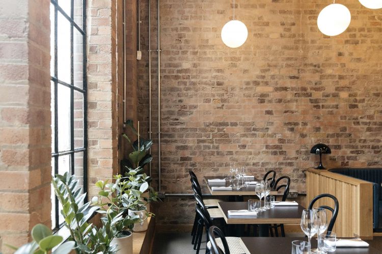 A dining area with natural light from large windows, featuring exposed brick walls, hanging globe lights, and tables set with glassware and green potted plants.