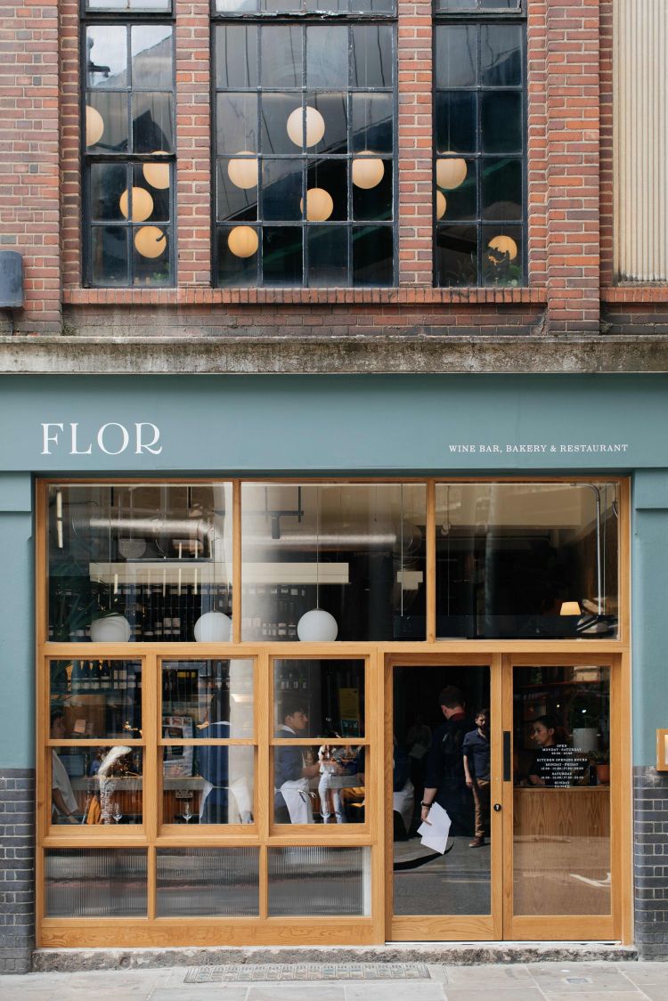 The exterior of FLOR restaurant, showcasing its glass facade with wooden frames, interior lighting, and reflections of hanging globe lights in the windows, set against a red brick building.