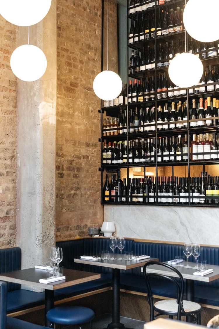 The interior of a restaurant with a wine rack wall, spherical pendant lights, and dining tables with blue upholstered bench seating against an exposed brick wall.