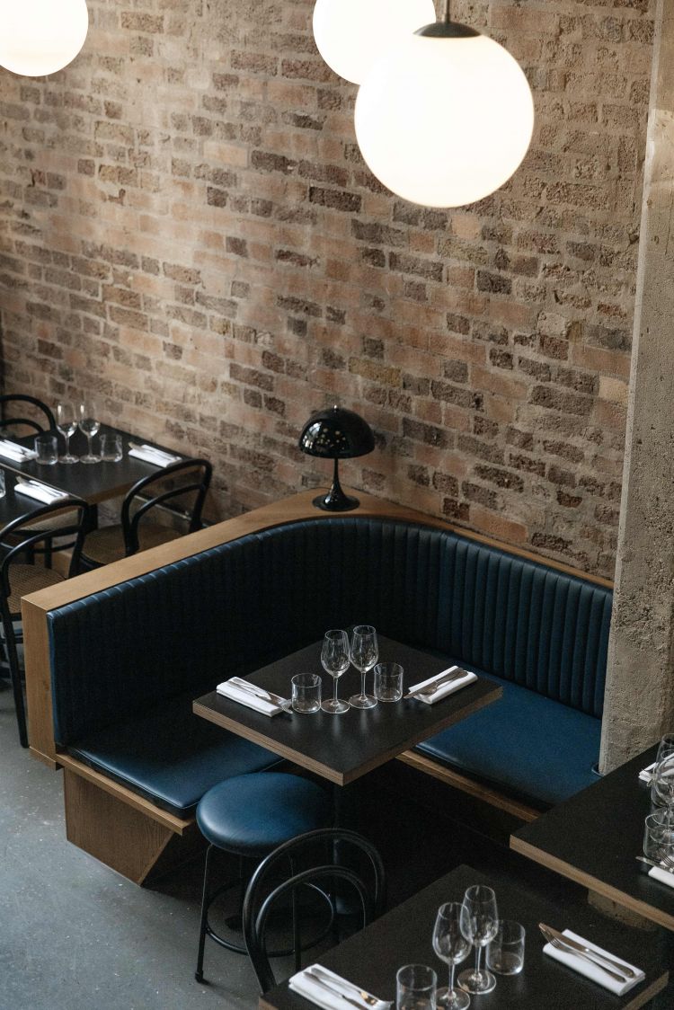 A restaurant corner with a blue upholstered booth, wooden tables set with wine glasses, and hanging globe lights against a backdrop of an exposed brick wall.