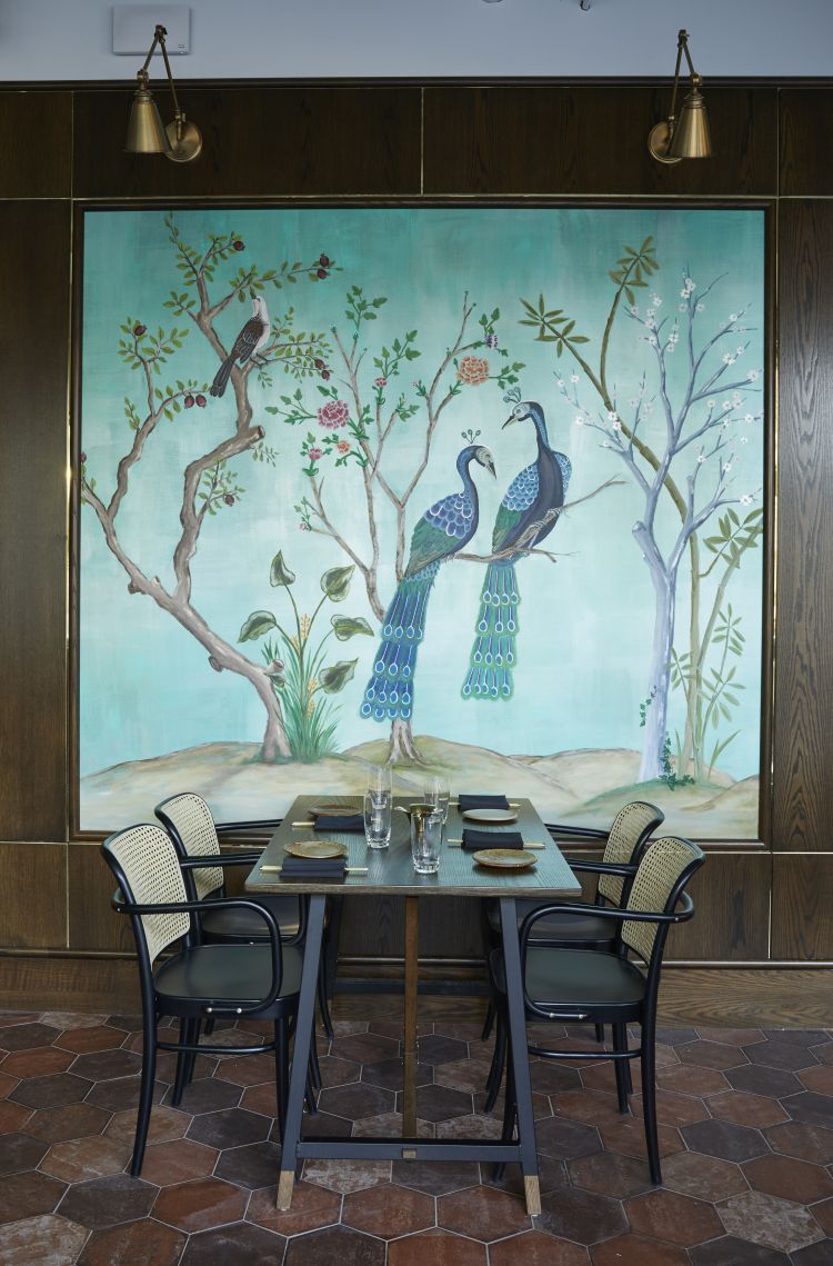 A dining table set against a large, decorative wall painting of two peacocks amidst a floral landscape, with elegant wall-mounted lamps above and terracotta-tiled flooring.