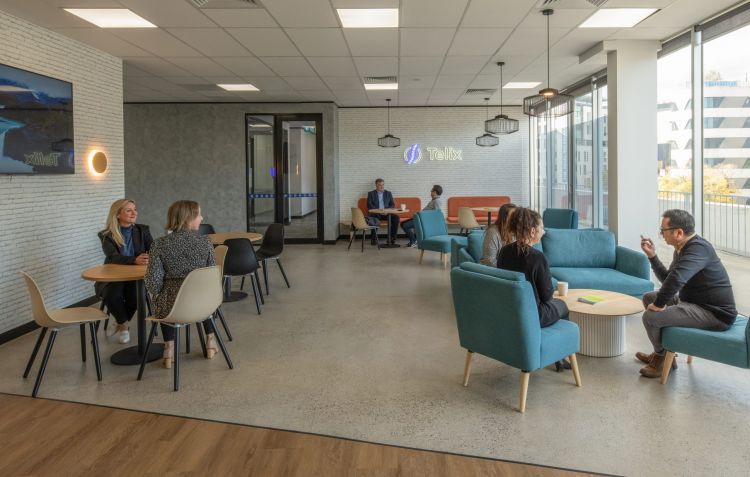 A modern office lounge with employees engaged in conversation, featuring contemporary furniture, large windows, and a logo of Telix Pharmaceuticals on the wall.