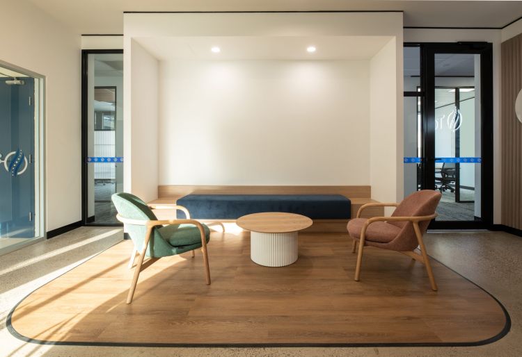 A simple office seating area with a wooden bench, round table, and two armchairs, illuminated by natural light from the side windows.