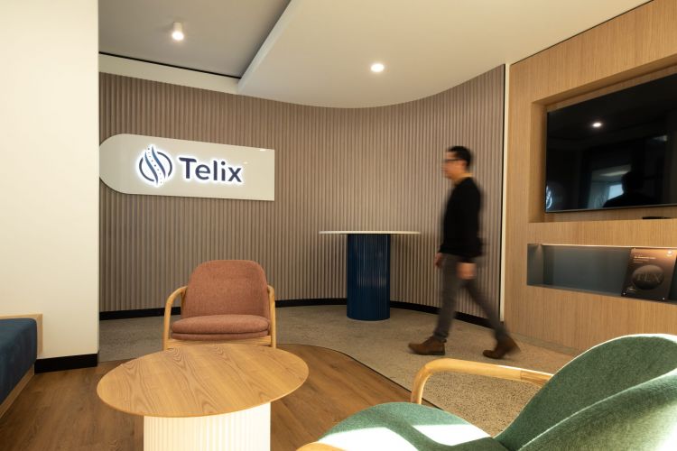 An office interior with a person walking past, a round table with chairs, and a lit-up Telix Pharmaceuticals logo on the wall.