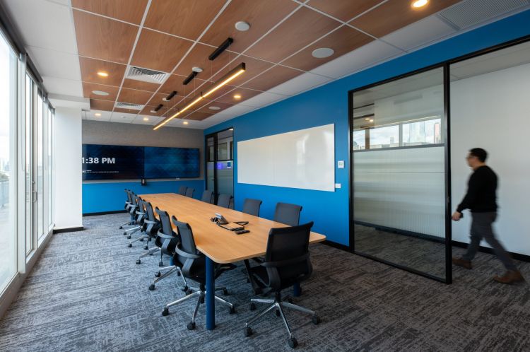 "A spacious conference room with a long table, chairs, and a digital screen on the wall, with a person walking by. 