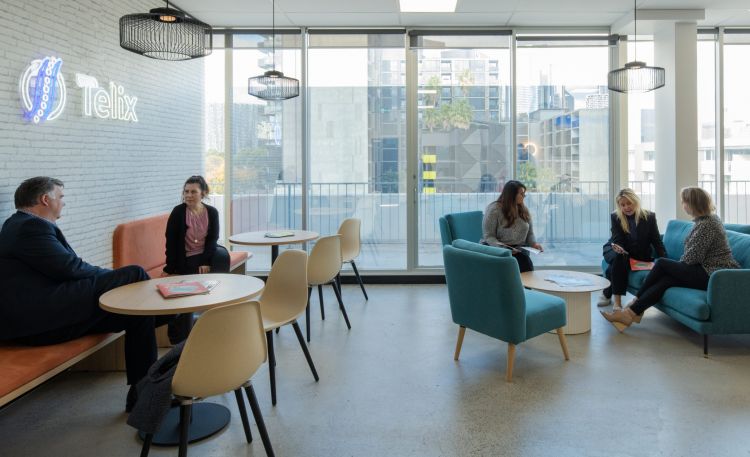 A well-lit office lounge area with employees having discussions, comfortable seating, large windows, and the Telix logo on the wall.