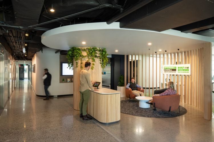 An office reception area with a curved desk, seating, hanging plants, and people in conversation, featuring the McConnell Dowell | Built Environs logo.