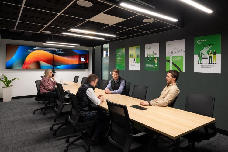 Four people in a modern office meeting room with green-themed motivational posters on the wall.
