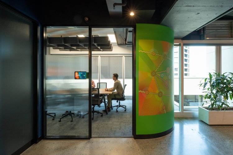 View into a modern office space through glass doors, showing people at a meeting table and a colorful abstract art column to the right.
