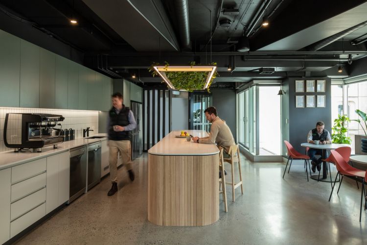 Office kitchen with a long wooden island and bar stools, modern coffee machine, and a round table with Pebble chairs.