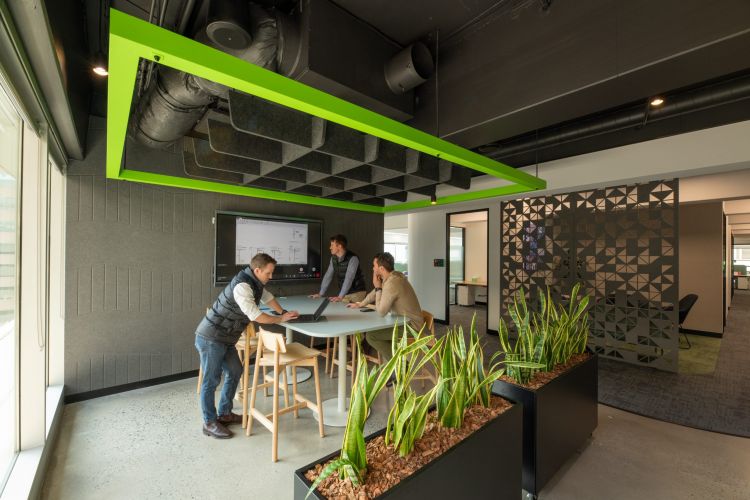 "A collaborative office space with a high wooden table, Andi bar stools, and a large screen on the wall, accented with greenery and decorative partitions.